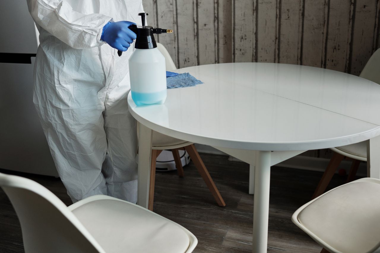 hero-img-01 Person in protective suit disinfecting a white table indoors with a sprayer.